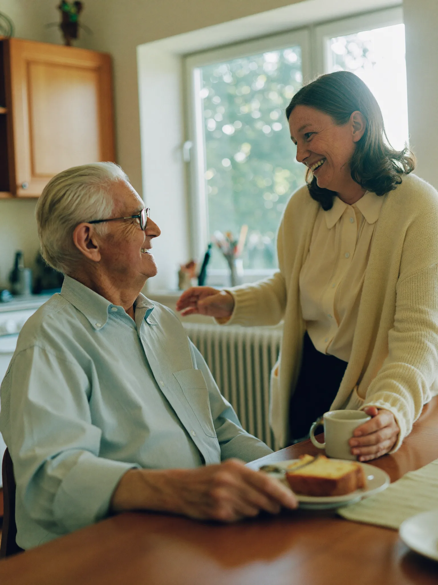 Betreuungskraft und Senior beim Kaffee am Küchentisch, beide lachen